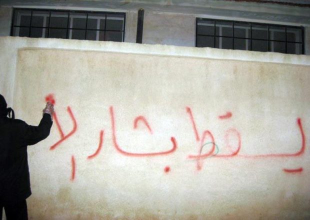 A person spraypainting graffiti on a wall in Arabic. Translation: Down with Bashar