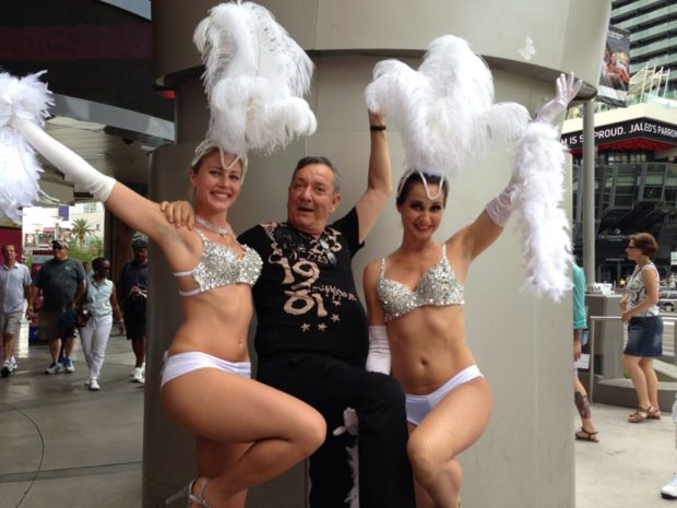 Photo of a senior man flanked by two Vegas showgirls with large feathered hats. They are smiling and each holding one arm in the air.