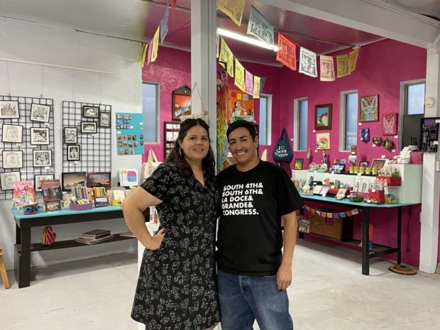Mel Dominguez and Melissa Brown Dominguez stand in their gallery with a colorful wall, papel picado, and community art hanging in the background