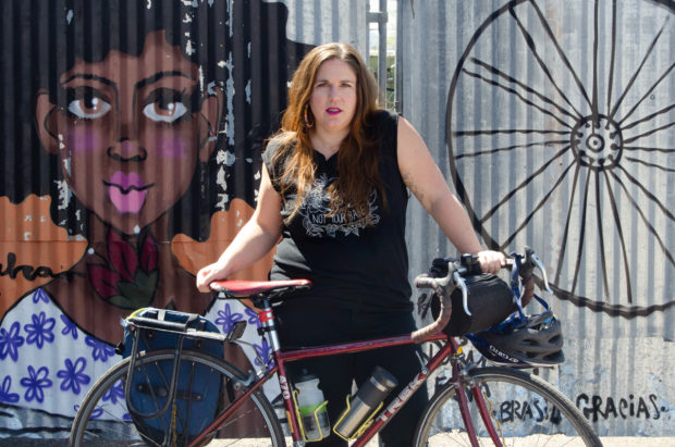 Kristin Mcray stands confidently holding her burgundy bike. She wears her light brown hair down, red lipstick, and black graphic thick tank top.