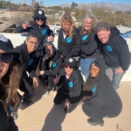 9 KXCI Staff members smiling in the sun wearing KXCI black hoodies with blue logs and bucket hats.