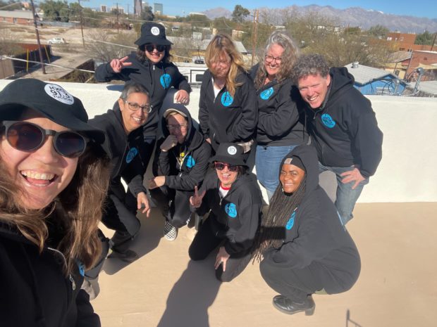 9 KXCI Staff members smiling in the sun wearing KXCI black hoodies with blue logs and bucket hats.