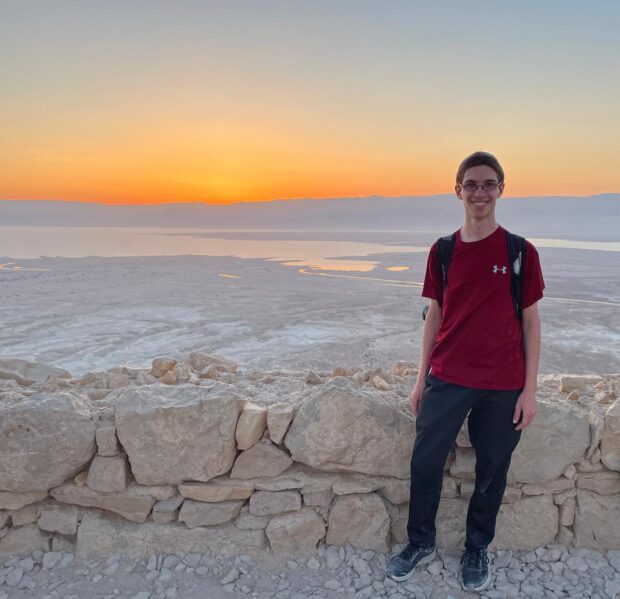 Young man with a back pack and red shirt standing at sunset near a rock wall and a spectacular mysterious landscape