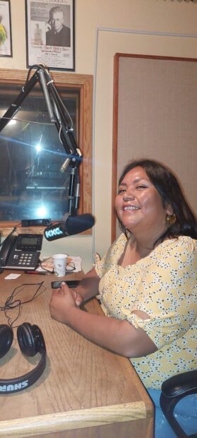Amy Juan is seated at The microphone in studio 2B at KXCI Community Radio. She is smiling and there is a phone near her.