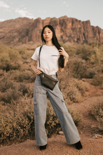 Young woman with long dark hair swept to the side standing against a landscape of  mountains and desert plant life, holding a handbag and looking like a fashion model with a serious facial expression.
