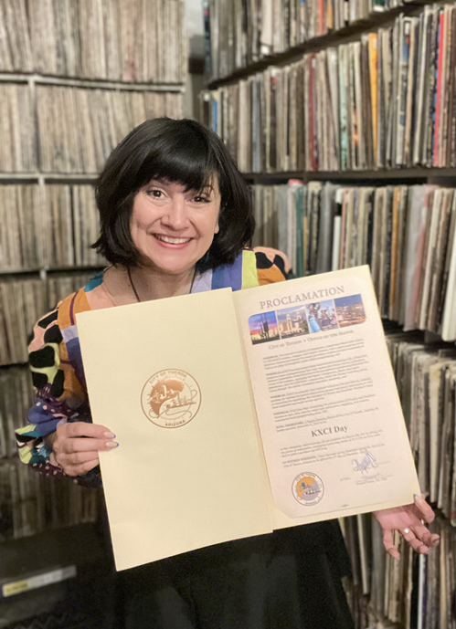 Bridgitte Thum holds KXCI Day Mayoral Proclamation in front of albums
