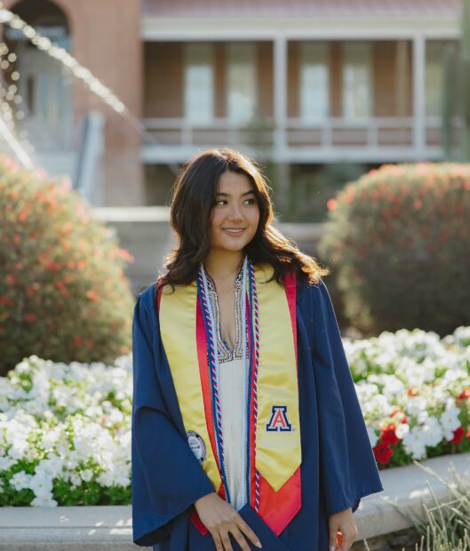 Young woman in graduation gown on campus the sun is shining down and highlighting rerd golden tones in her brown long hair. She is wearing a special sash with a logo on it representing the U of A