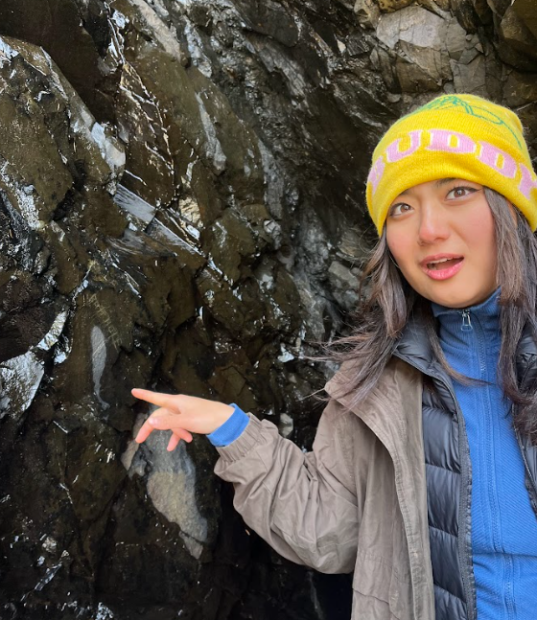 A young wonam is wearing a yellow sock hat and pointing to a rock wall with a look of astonishment.