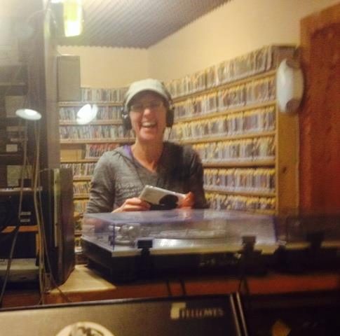 Susie B. has a cap and headphones and she is smiling in the KXCI studios with cds in the background.